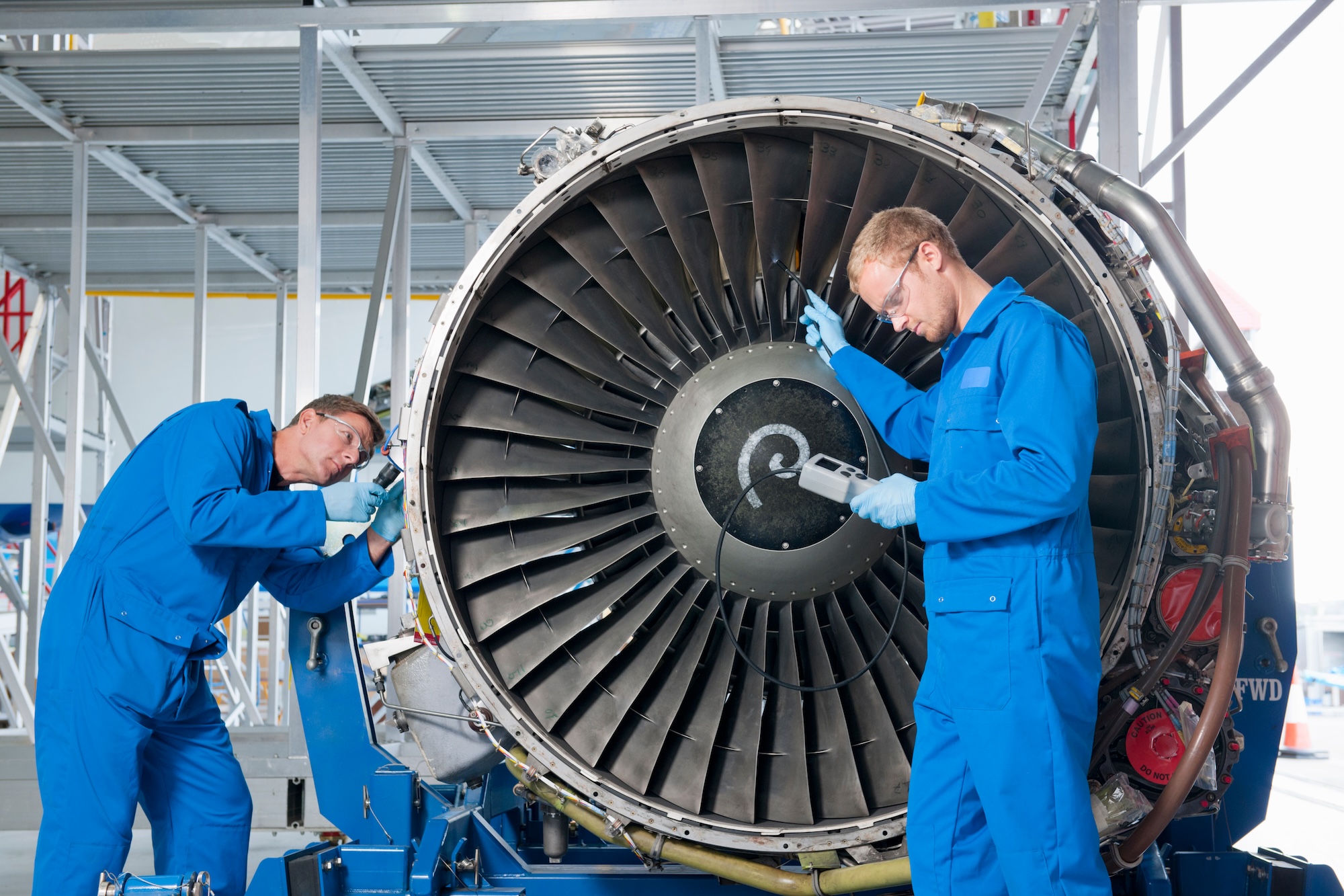 Two aerospace engineers in blue uniforms inspecting and maintaining aircraft turbine blades in modern aerospace manufacturing facility