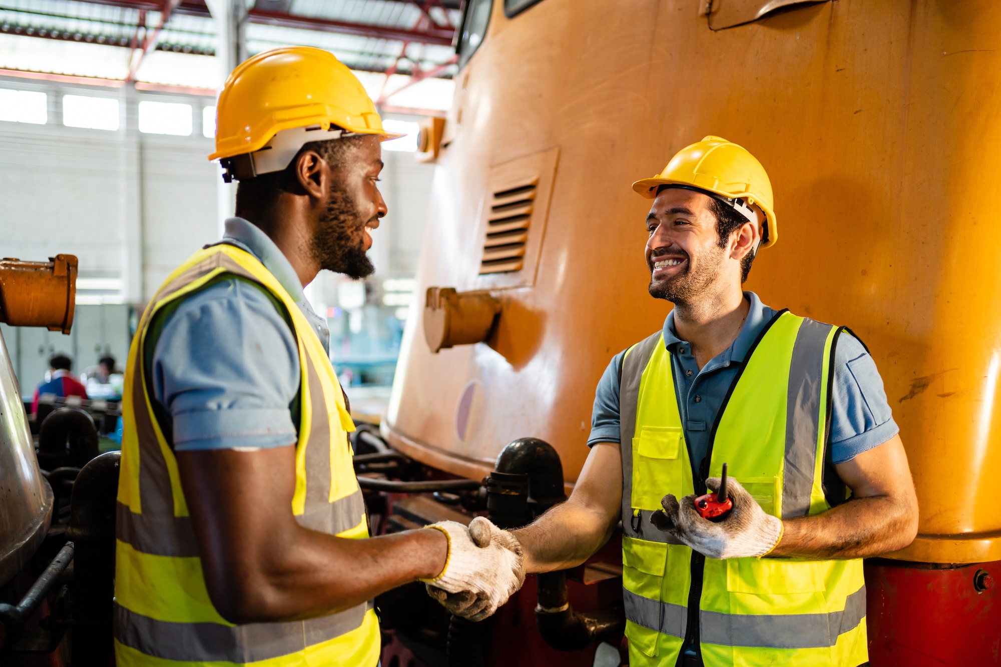 Two subcontractor professionals in hard hats and safety vests shaking hands in manufacturing facility with industrial equipment
